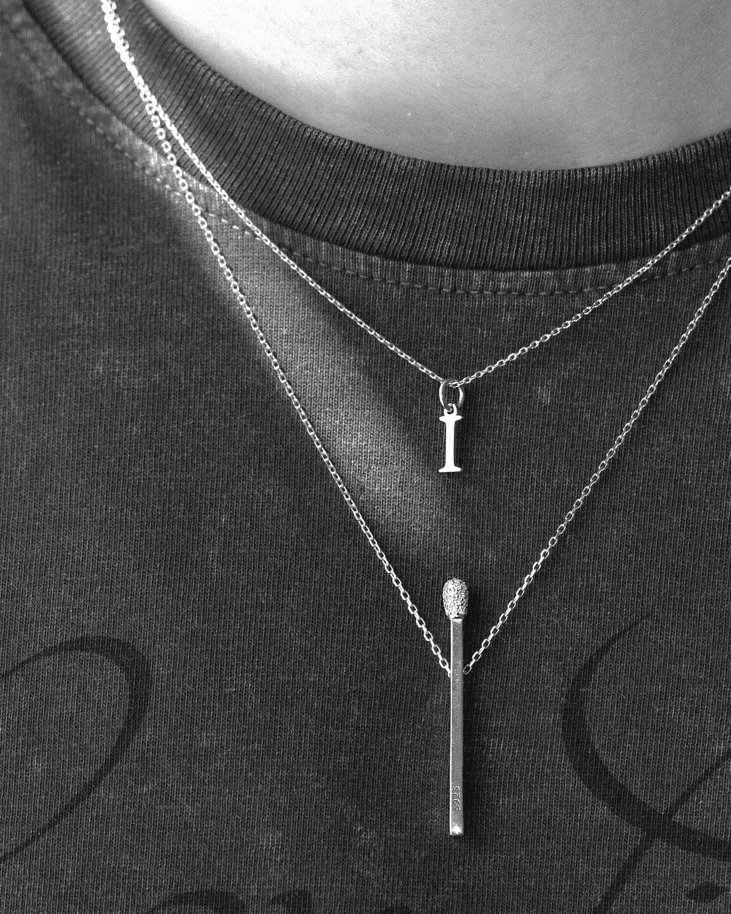 Two necklaces with a letter 'I' pendant and a cylindrical pendant on a black shirt.