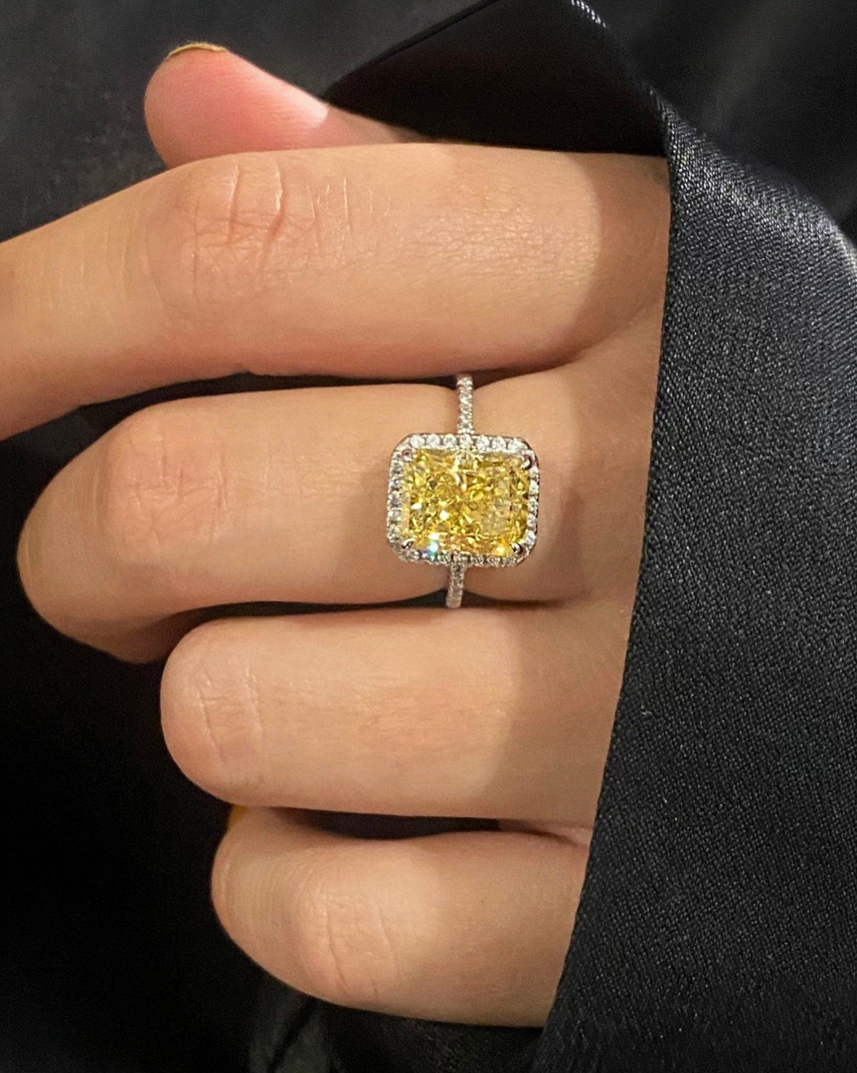 A close-up of a hand wearing a silver ring with a cushion-cut yellow center stone and a halo of small clear stones, set against a dark black fabric background.